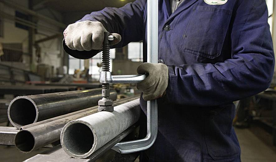 Ein Arbeiter in Schutzkleidung bearbeitet Metallrohre mit einer großen Zwinge in einer Werkstatt. Fokus liegt auf den Rohren und dem Werkzeug.