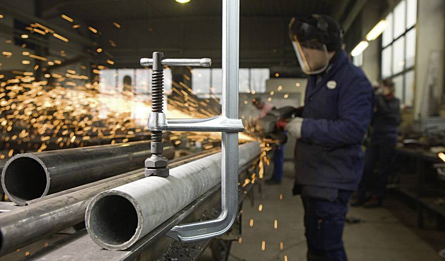Ein Arbeiter in einer Metallwerkstatt schneidet ein Metallrohr mit einem Winkelschleifer, Funken fliegen in alle Richtungen.