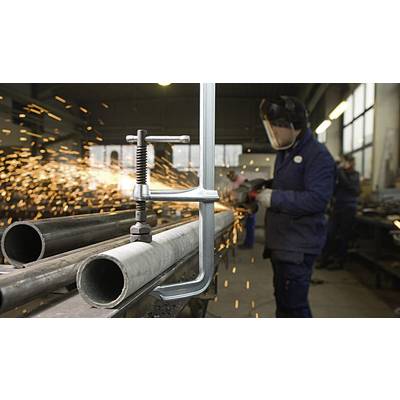 Arbeiter in Werkstatt schleift Metallrohr, Funken fliegen. Im Vordergrund sind zwei große Stahlrohre und eine Schraubzwinge sichtbar.