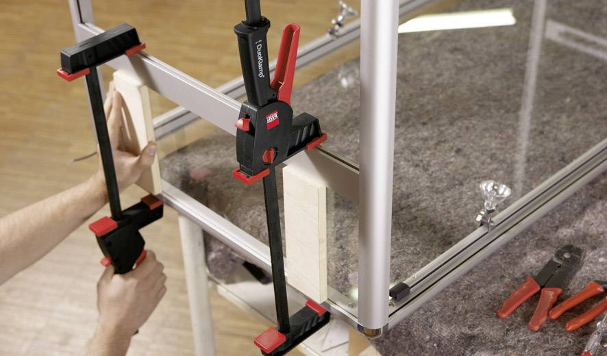 Zwei Hände nutzen Schraubzwingen, um Holzlatten an einem Metallrahmen zu fixieren. Auf einem Tisch liegen Werkzeuge.
