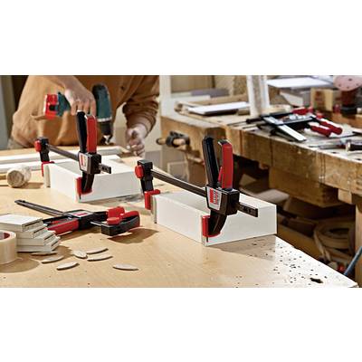 Eine Person arbeitet an einer Holzbank und bohrt in Holzstücke, die mit roten Klemmen fixiert sind. Holzspäne liegen auf dem Tisch.