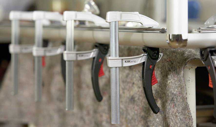 Mehrere Klemmen halten Stoff an einem Tisch fest, wahrscheinlich in einer Werkstattumgebung. Klemmen sind silber mit schwarzen Griffen.
