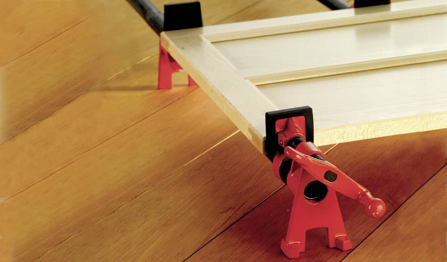 Eine rote Klemme hält ein hölzernes Brett auf einem lackierten Holzboden fest. Die Klemme fixiert das Brett vermutlich für handwerkliche Arbeiten.