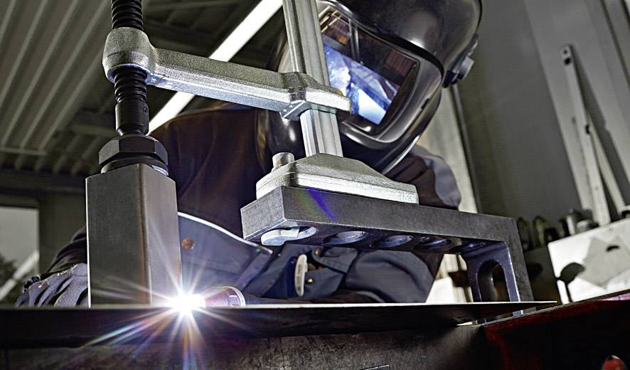 Eine Person in Schutzkleidung schweißt ein Metallstück in einer Werkstatt. Funken fliegen, während das Schweißgerät arbeitet.
