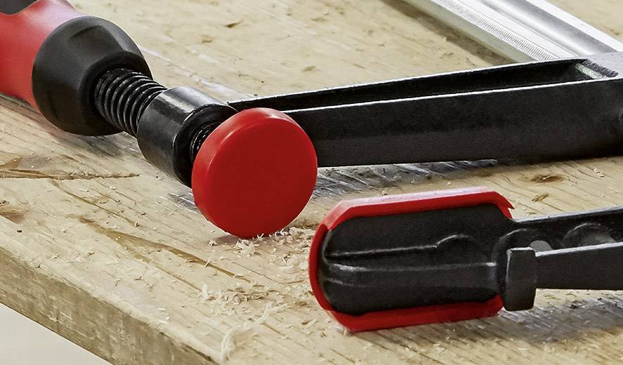 Ein Schraubzwinge auf einer Holzoberfläche. Die Zwinge hat rote Kunststoff-Enden, die ein Verrutschen verhindern. Holzspäne sind sichtbar.