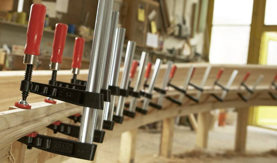 Holzwerkstatt mit mehreren Klemmen, die gebogene Holzstücke fixieren. Hintergrund zeigt Werkzeuge und Regale.