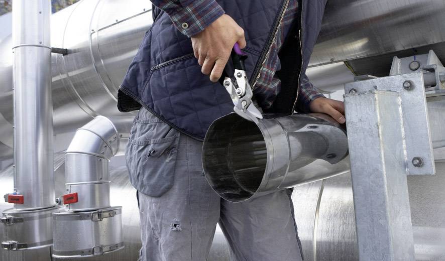Person schneidet ein Metallrohr mit einer Metallschere auf einer Baustelle. Im Hintergrund sind weitere Metallrohre sichtbar.