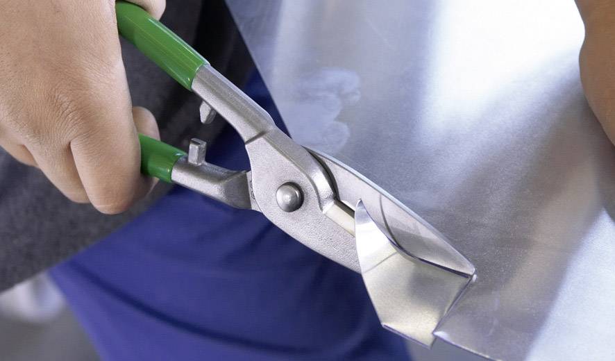 Eine Person schneidet ein Stück Metall mit einer grünen Metallschere. Die Metallschere ist in Nahaufnahme zu sehen.