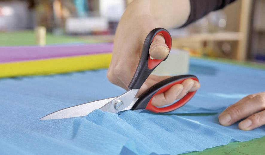 Eine Hand schneidet mit einer Schere blauen Stoff auf einem Tisch. Im Hintergrund sind bunte Stoffe und Nähutensilien zu sehen.
