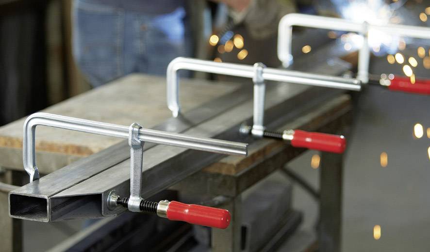 Metallverarbeitungsprozess: Mehrere Metallstücke werden mit roten Klemmen fixiert. Funken fliegen durch Schweißarbeiten im Hintergrund.