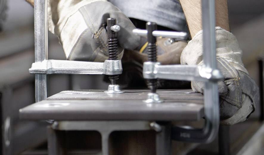Ein Handwerker mit Arbeitshandschuhen verwendet Metallzwingen, um ein Werkstück auf einem Tisch zu fixieren.