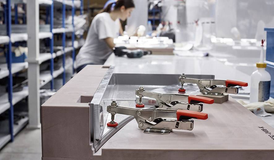 Eine Produktionshalle mit einem Tisch, auf dem verstellbare Klemmen ein Metallstück fixieren. Eine Person im Hintergrund arbeitet konzentriert.