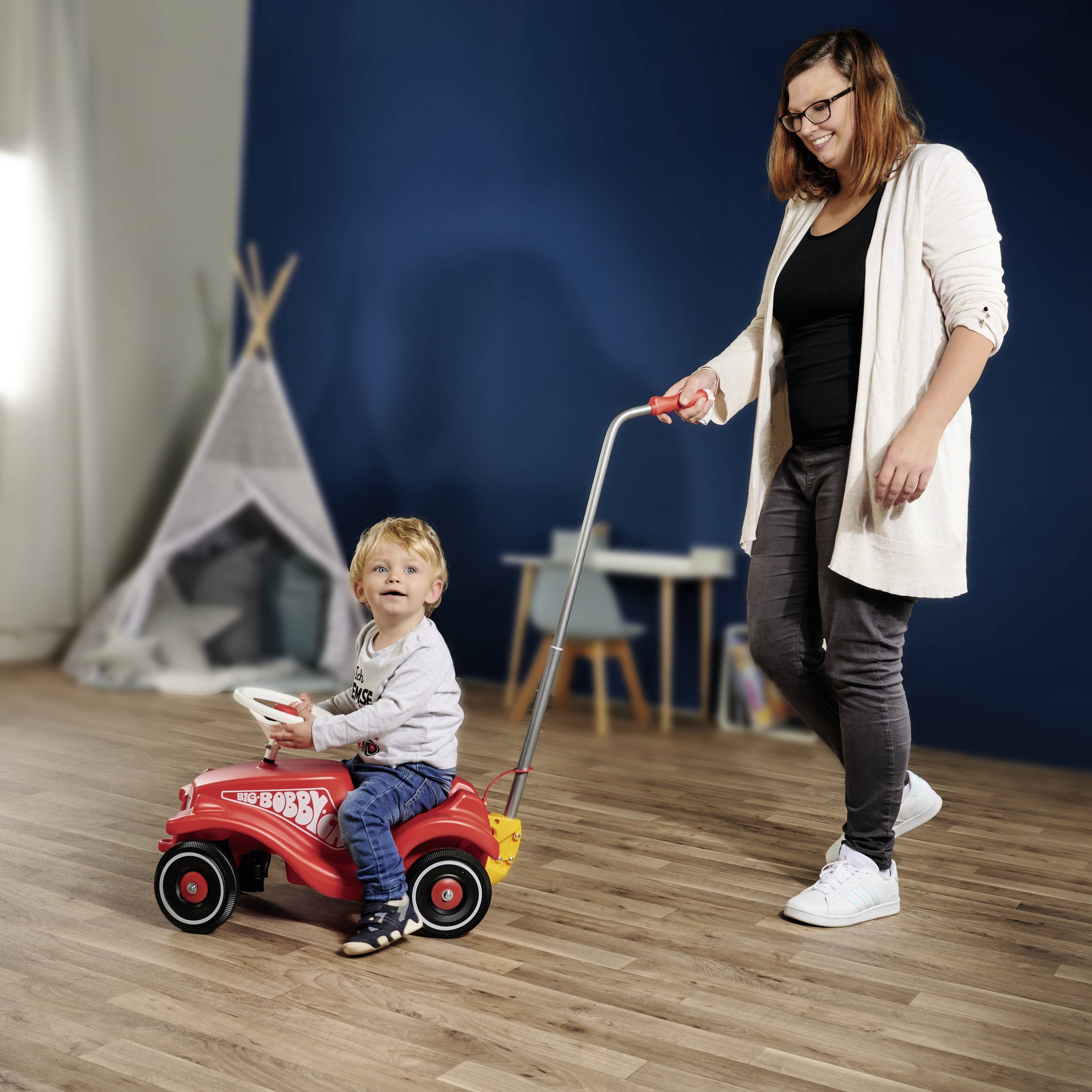 Eine Frau schiebt ein kleines Kind auf einem roten Spielauto durch einen Raum. Im Hintergrund ist ein Spielzelt zu sehen.