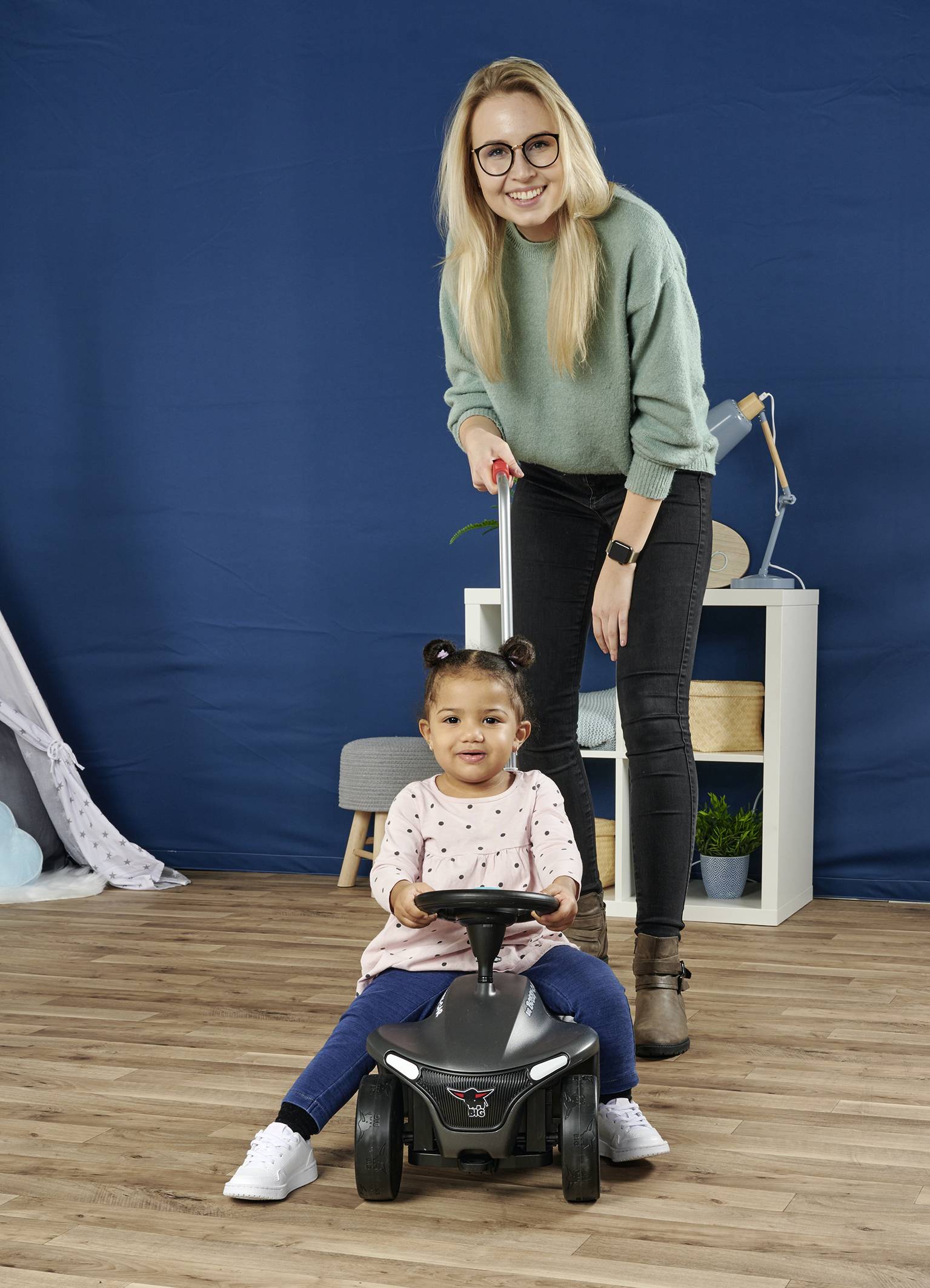 Eine Frau steht hinter einem Kleinkind, das auf einem kleinen Spielzeugauto sitzt. Sie lächeln in die Kamera.