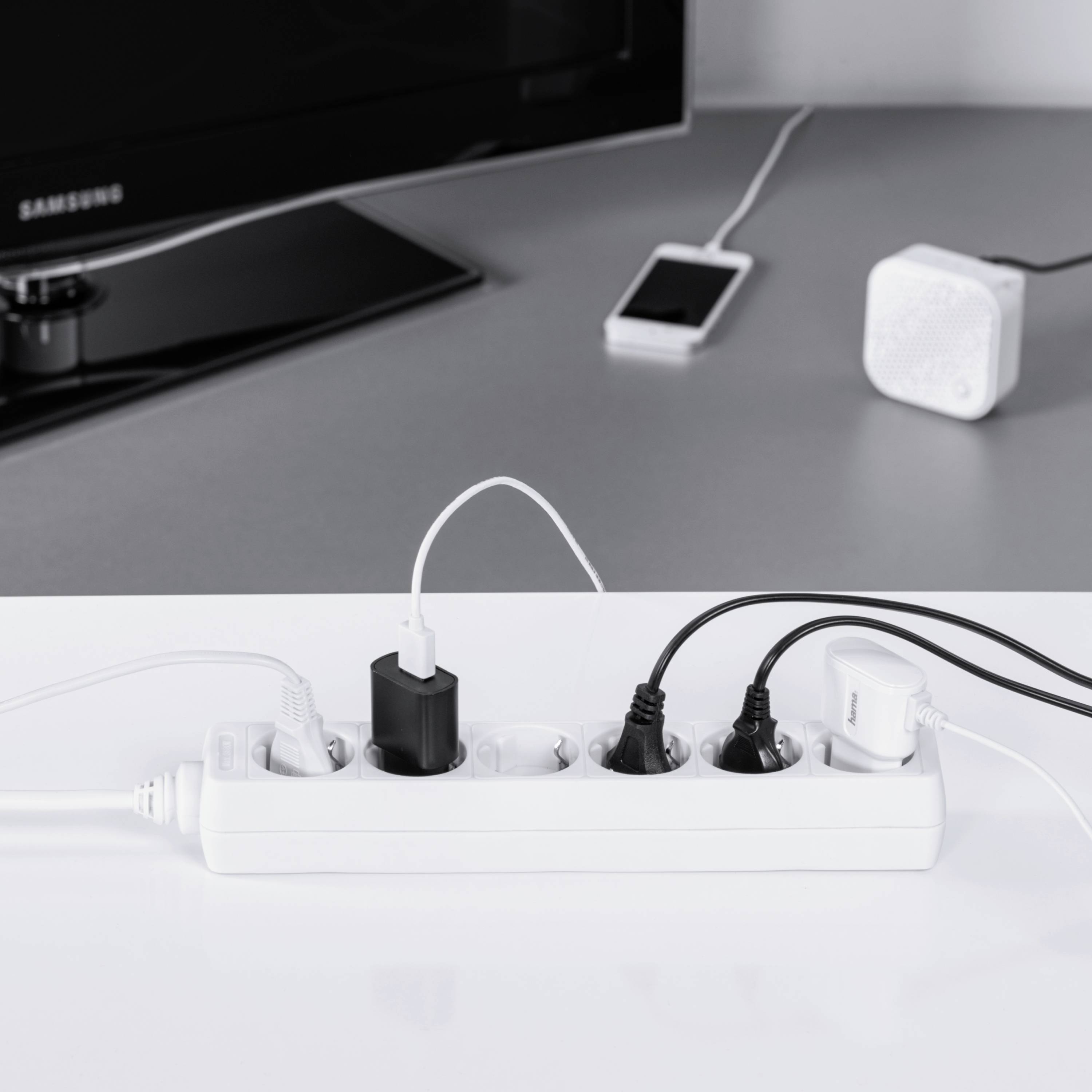Ein Steckdosenleiste mit mehreren angeschlossenen Geräten, darunter ein Smartphone-Ladegerät, vor einem Fernseher und einem Bluetooth-Lautsprecher.