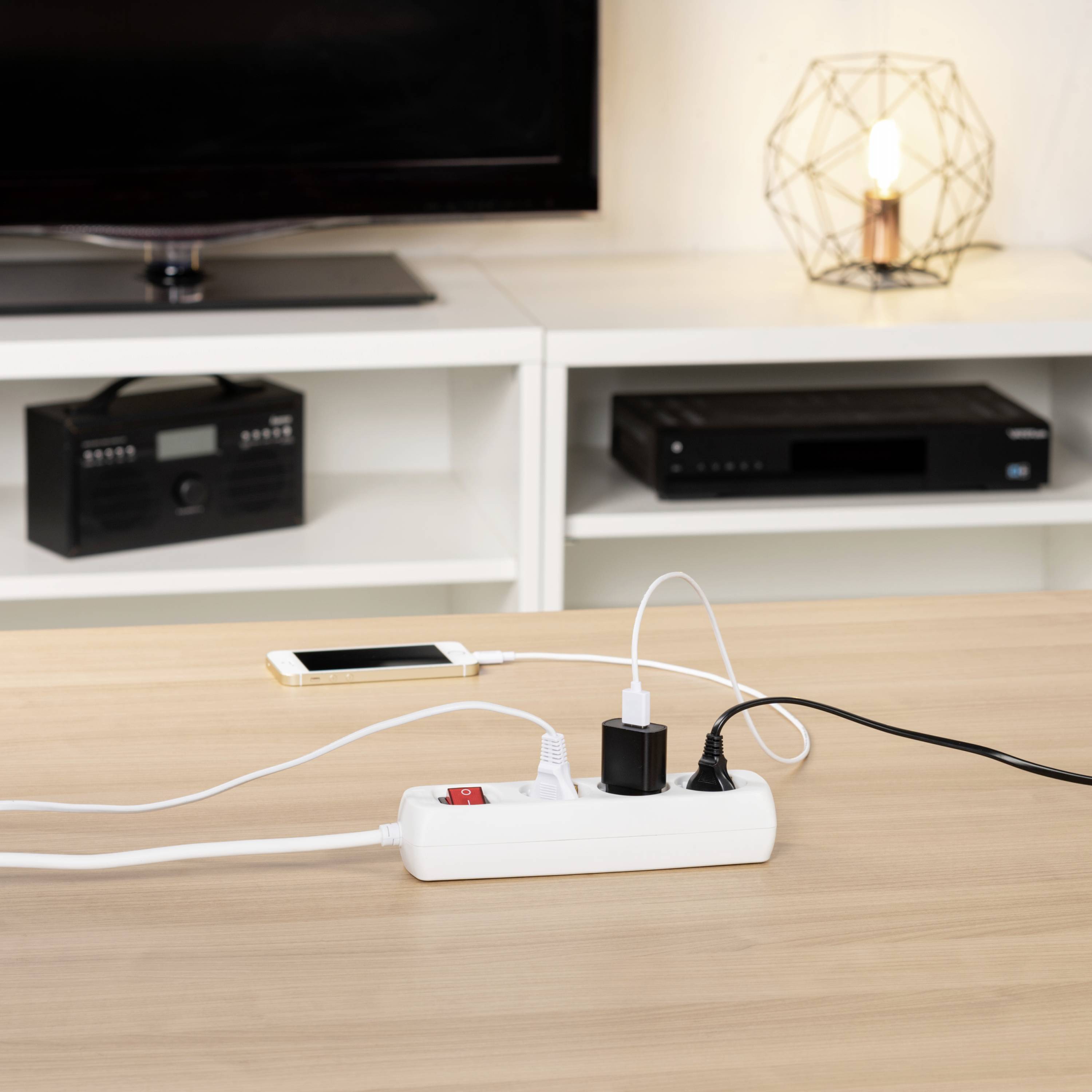Ein weißer Mehrfachstecker auf einem Holztisch, an den ein Smartphone und ein Ladegerät angeschlossen sind. Im Hintergrund ein Radiogerät.