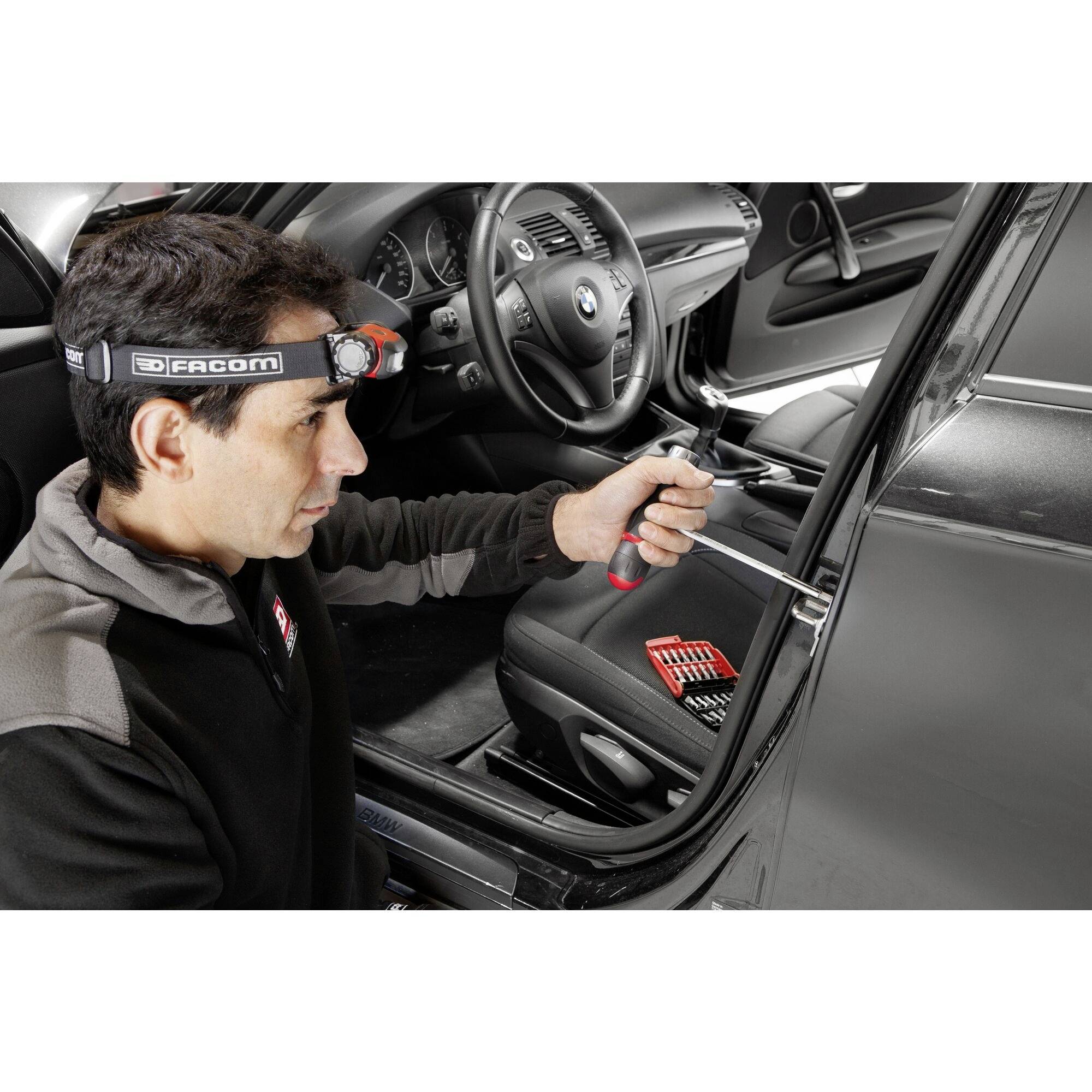 Ein Mann nutzt einen Schraubendreher zur Reparatur an der Autotür. Er trägt eine Stirnlampe und hat einen Werkzeugsatz in der Nähe.