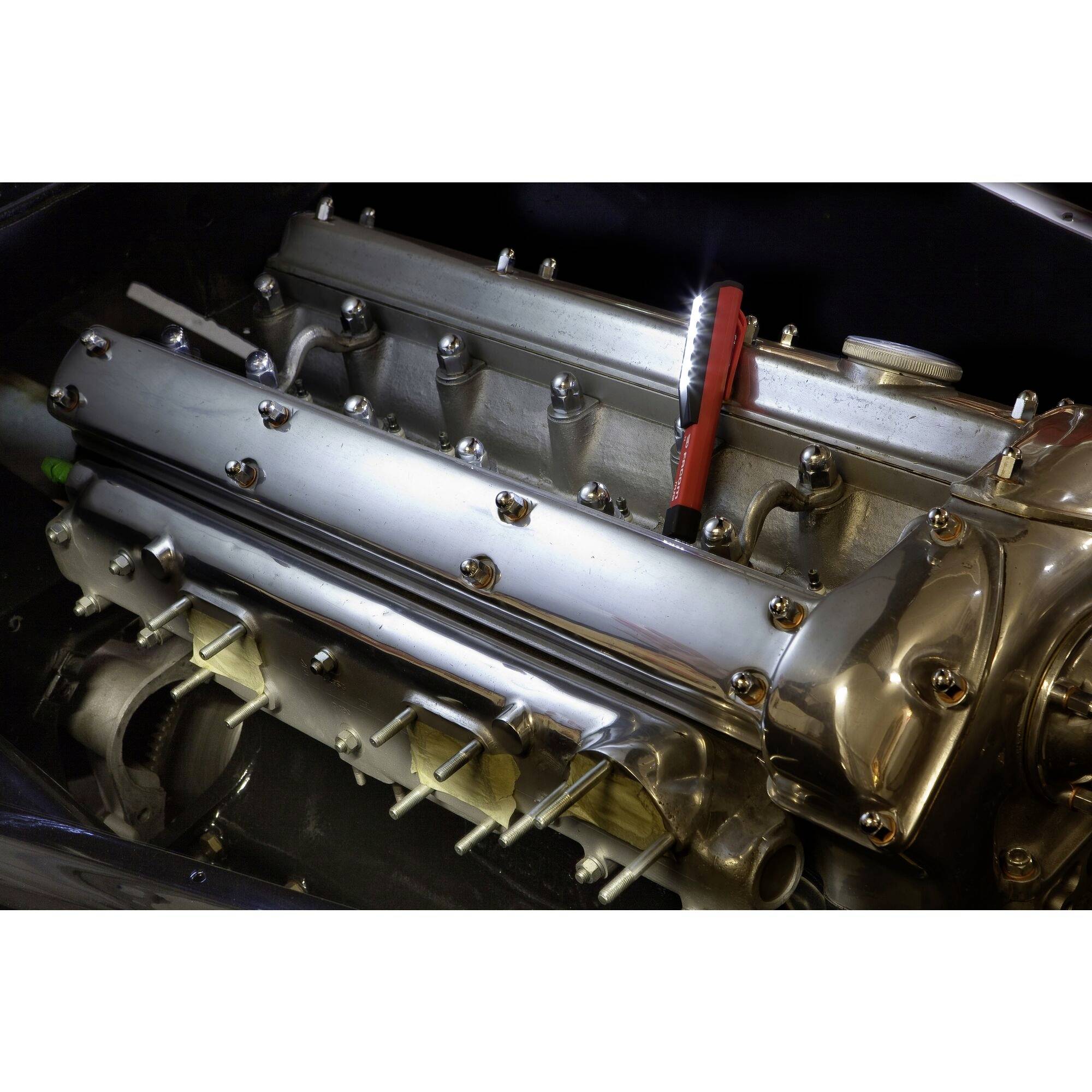 Ein glänzender, silberner Motorblock mit mehreren Zylindern und Bolzen ist zu sehen. Eine rote Zange liegt oben auf dem Motor.