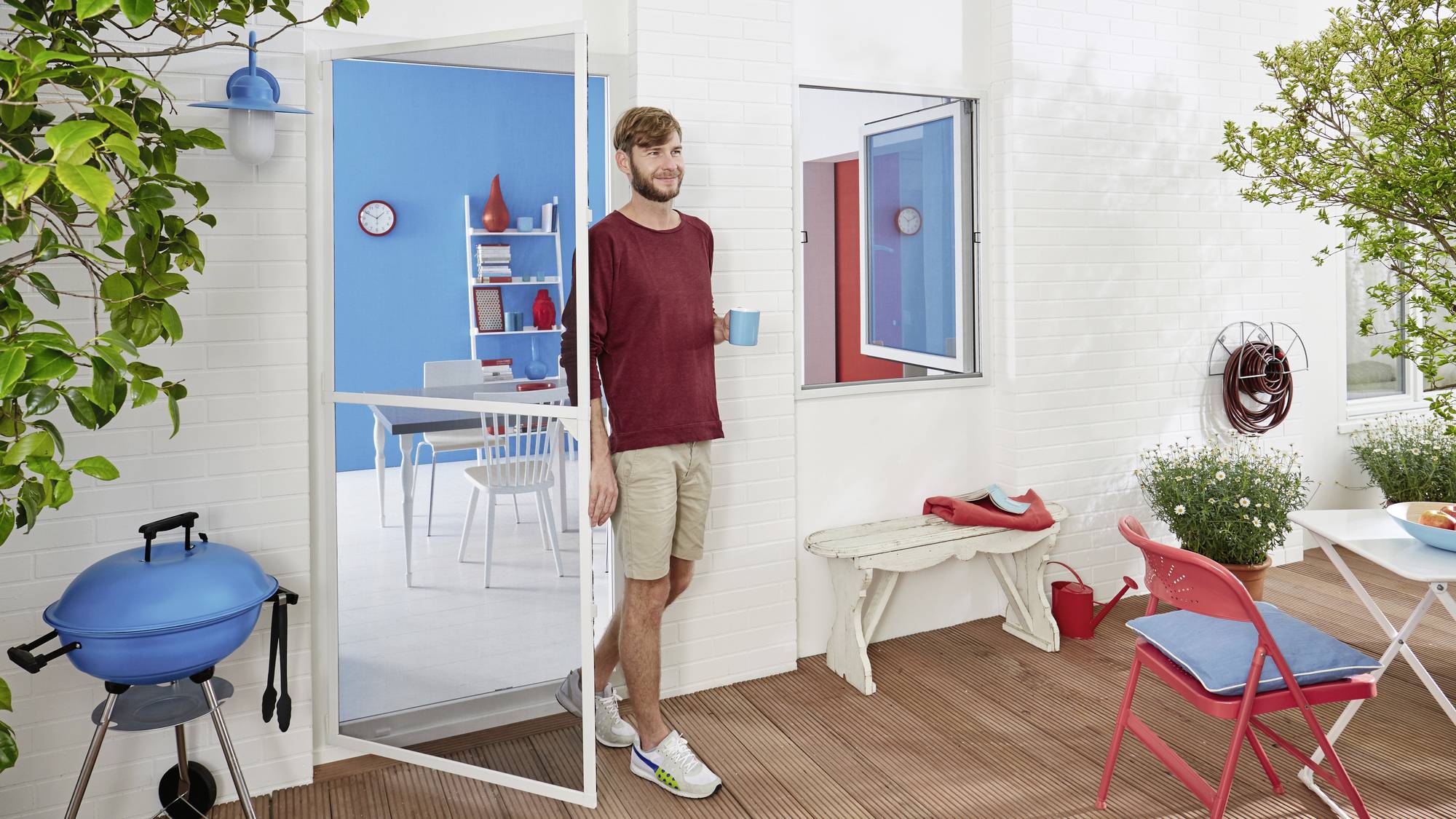 Ein Mann in Freizeitkleidung steht mit einer Tasse in der Hand in einer offenen Tür, die zu einem hellen, modern eingerichteten Raum führt.