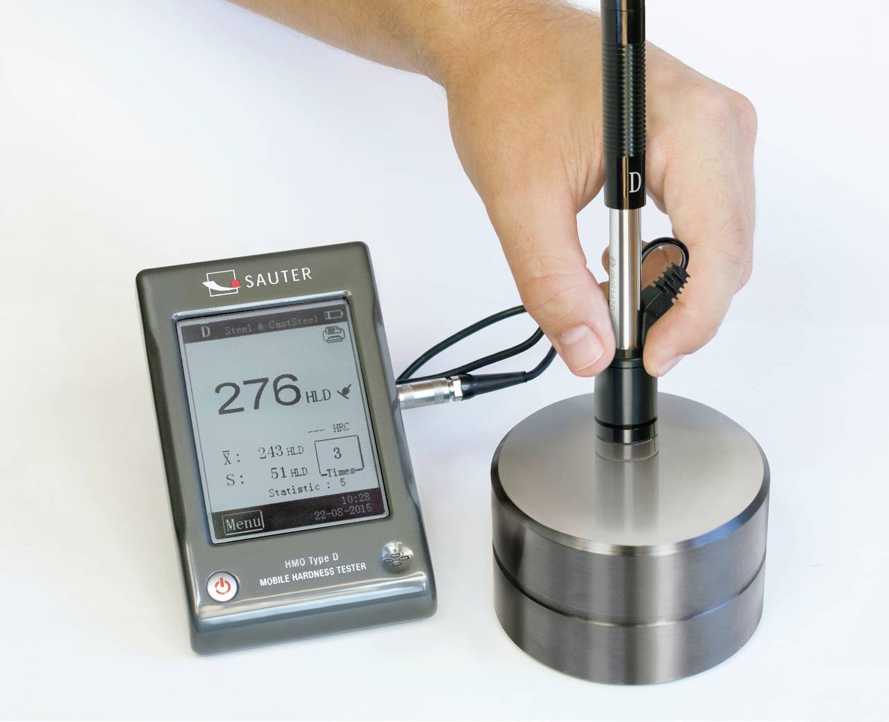 Eine Hand bedient ein digitales Härteprüfgerät, das auf einem Metallstück aufliegt. Das Display zeigt '276 HLD' als Messwert an.