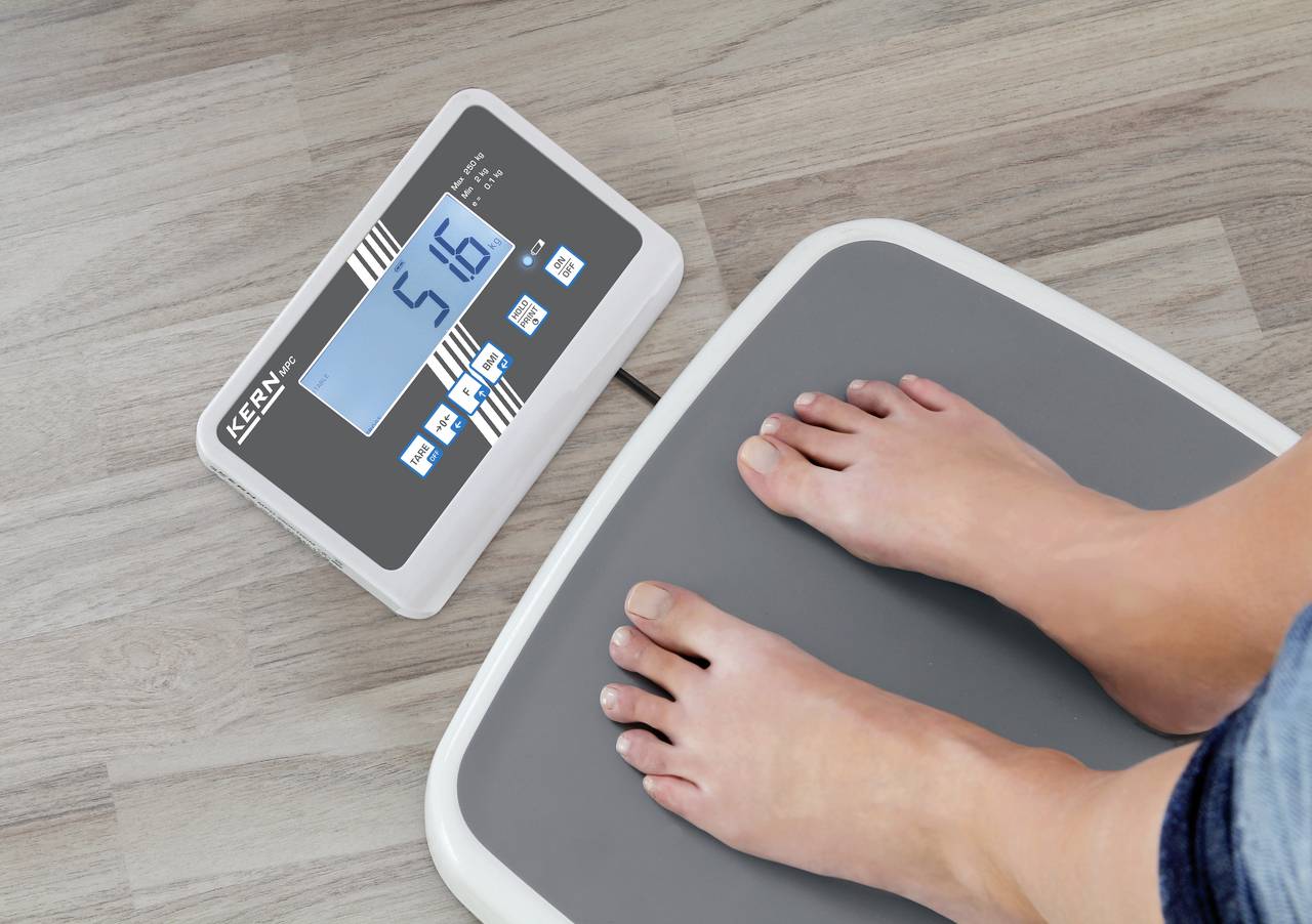 Person steht auf einer digitalen Waage, die '51,6' Kilogramm anzeigt. Die Waage liegt auf einem Holzboden.