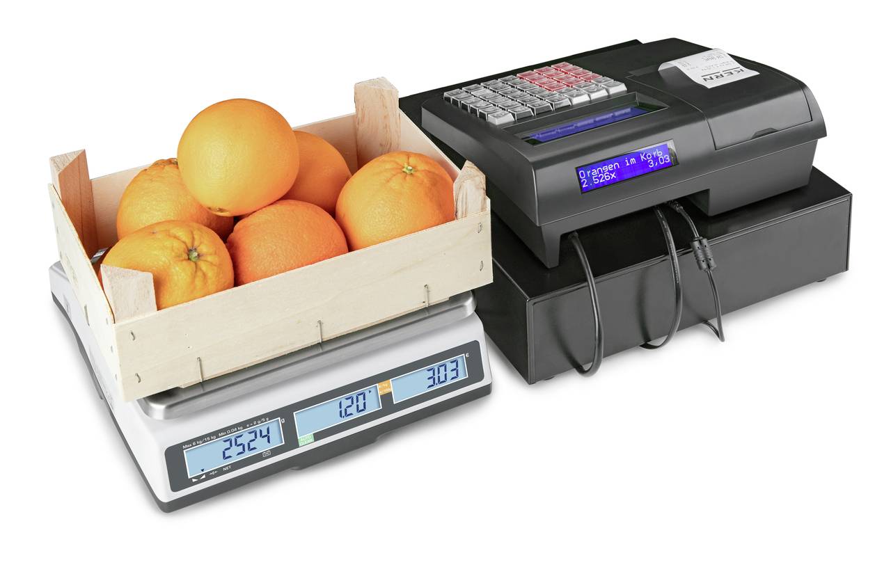 'Eine Holzkiste mit Orangen auf einer digitalen Waage, neben einer Registrierkasse mit sichtbarem Display und Quittungsausdruck.'