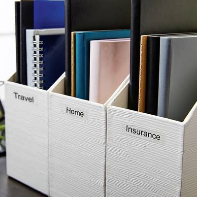 Drei weiße Zeitschriftenhalter auf einem Tisch, beschriftet mit 'Travel', 'Home' und 'Insurance'. Sie enthalten diverse Ordner und Dokumente.