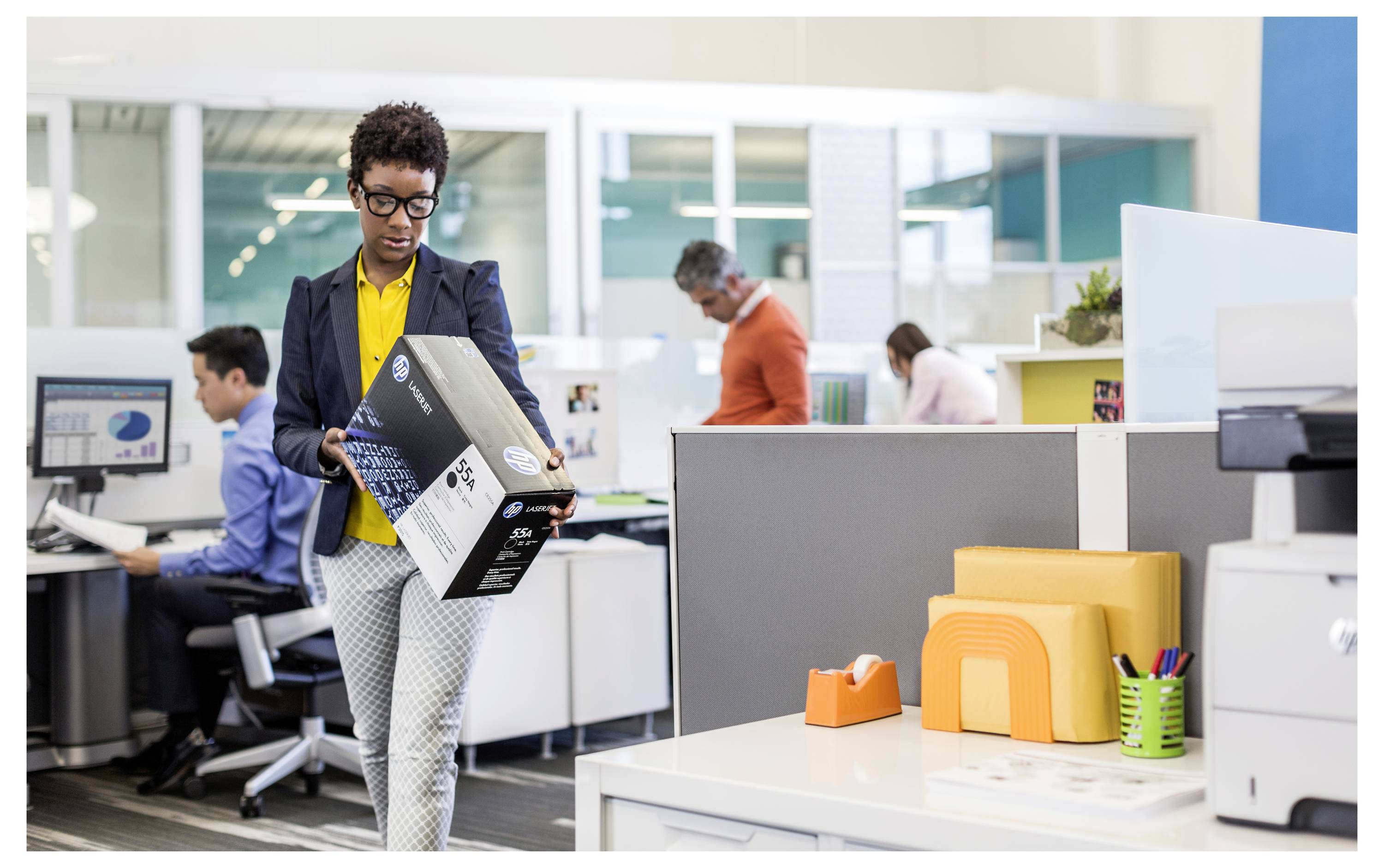 Büroangestellter, der einen verpackten Gegenstand durch ein modernes offenes Büro trägt, während Kollegen im Hintergrund an Schreibtischen arbeiten.