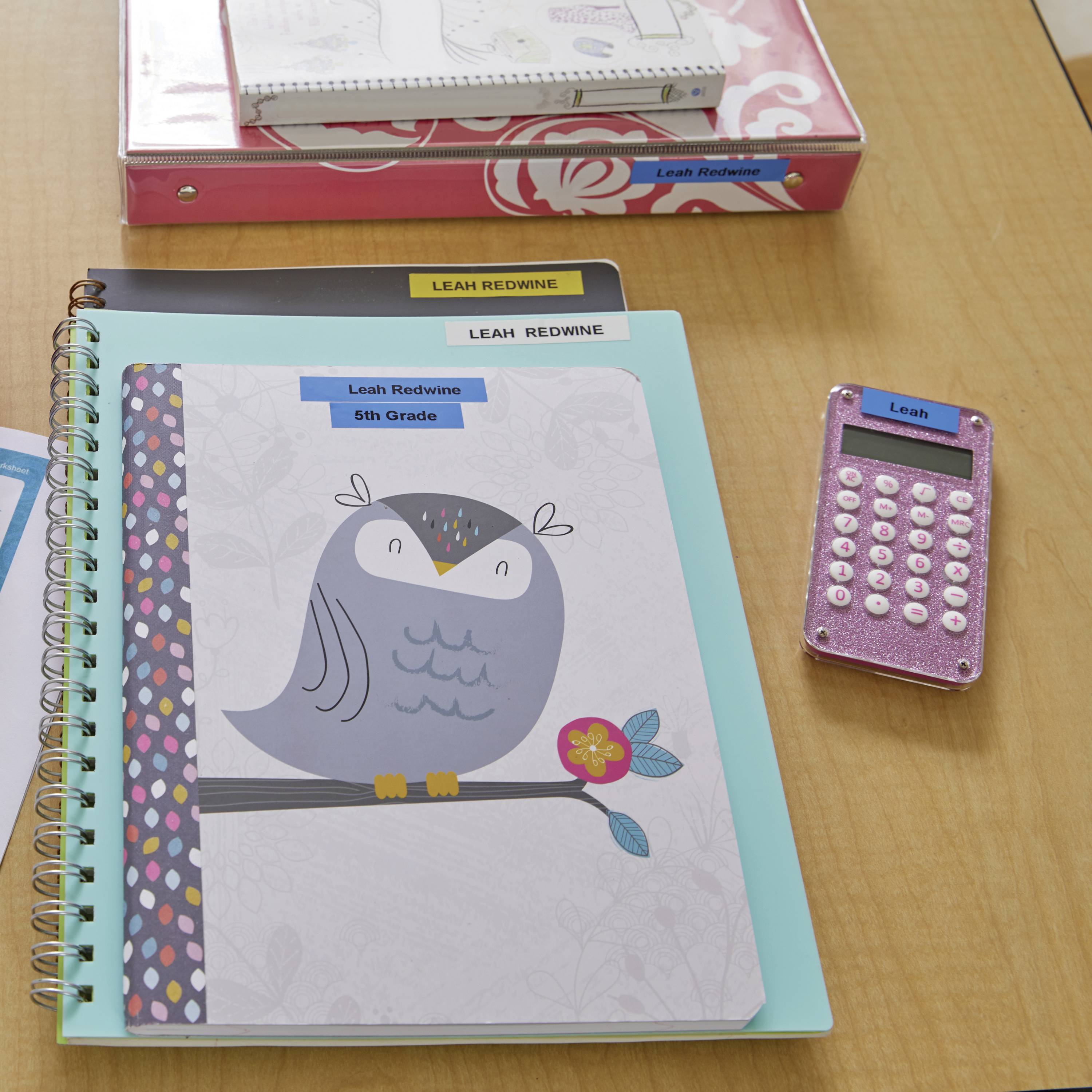 Auf einem Schreibtisch liegen Schulmaterialien: Ein Notizbuch mit Eulenmotiv, ein pinker Taschenrechner, und ein Ordner, alle mit dem Namen 'Leah Redwine' gekennzeichnet.