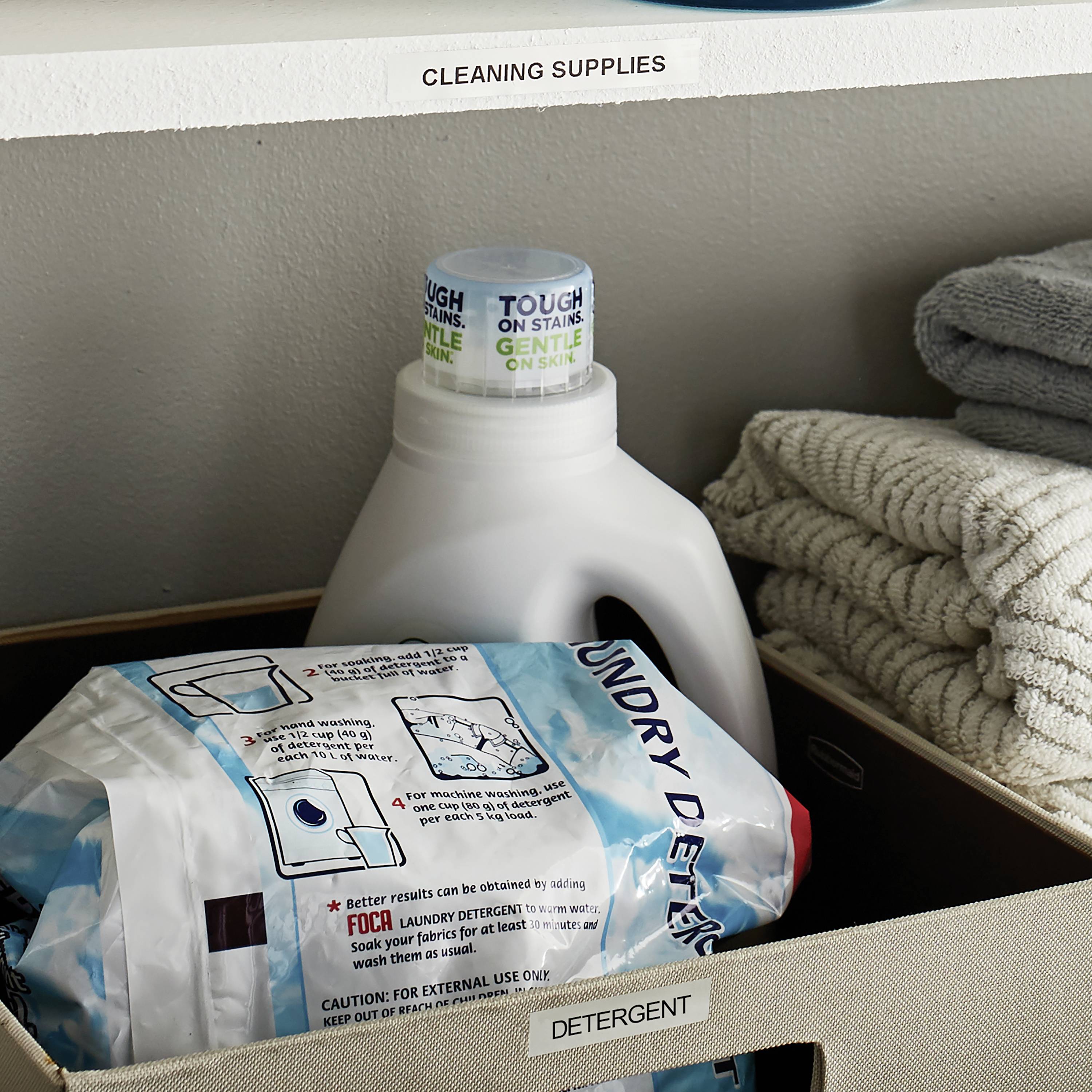 Reinigungsregal mit 'CLEANING SUPPLIES' Schild. Enthält Waschmittel in Flaschen und Tüten, daneben gefaltete Handtücher.