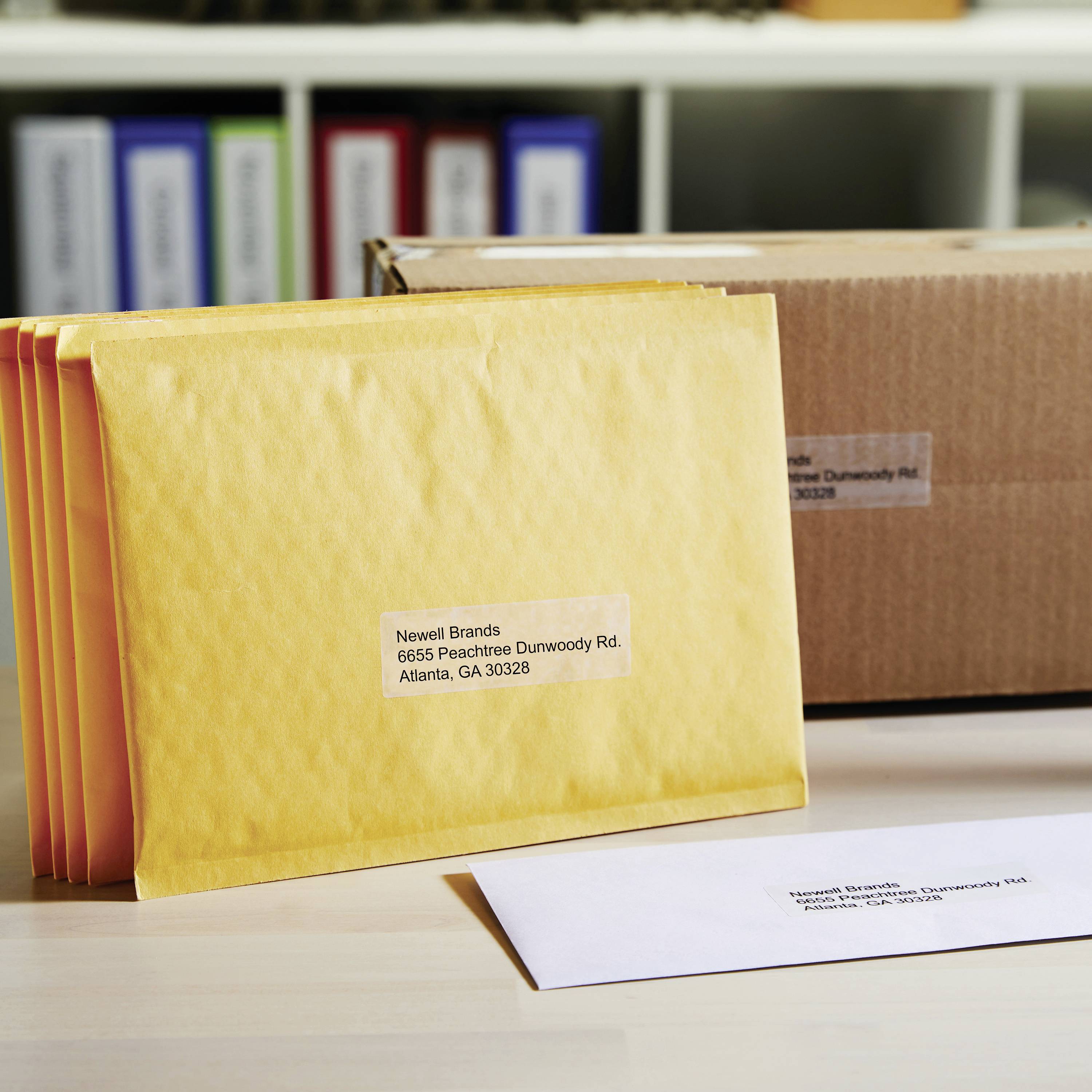 Ein Stapel gelber Versandtaschen und ein Briefumschlag mit der Adresse 'Newell Brands, 6655 Peachtree Dunwoody Rd, Atlanta, GA 30328'.