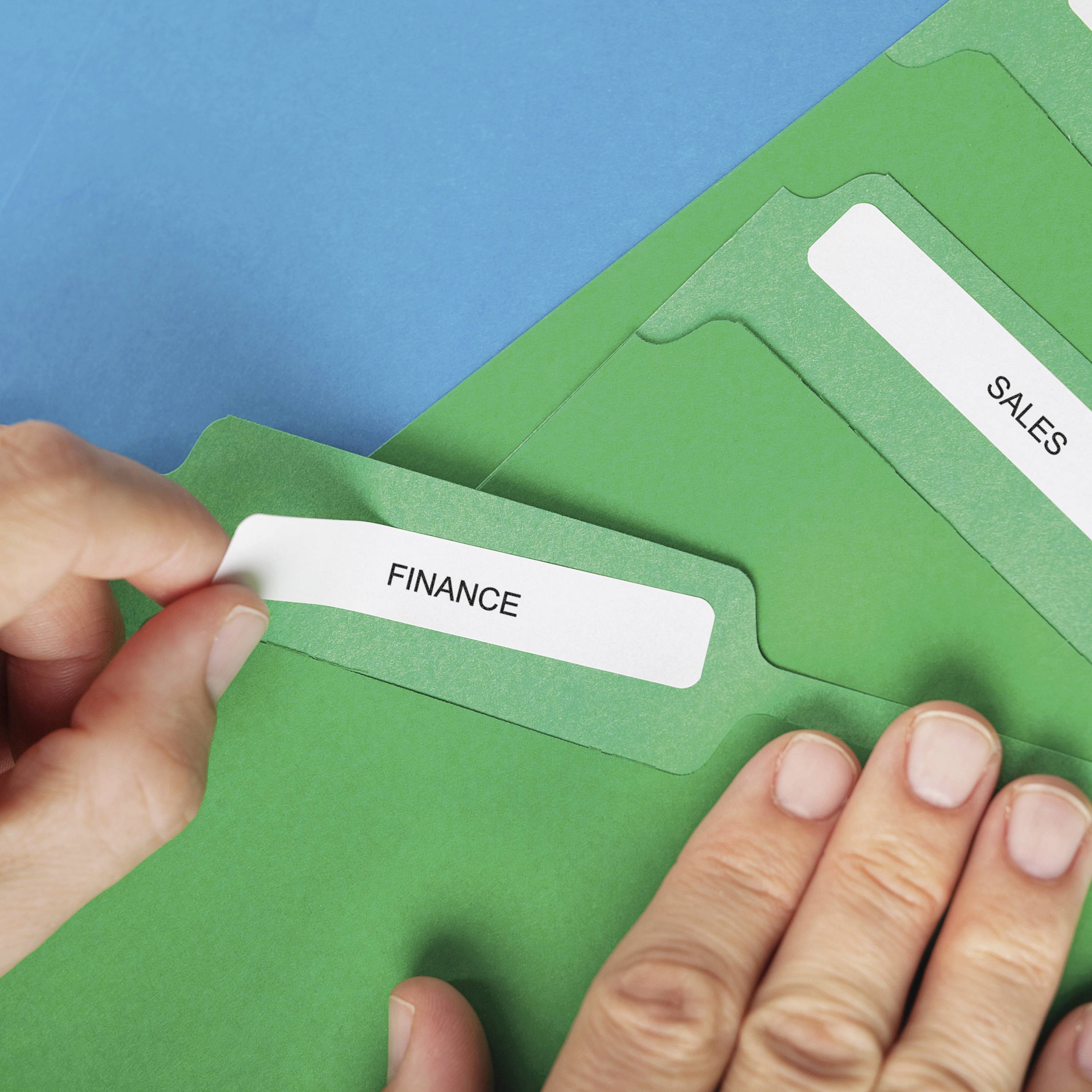 Hände sortieren grüne Ordner mit den Etiketten 'FINANCE' und 'SALES' auf blauem Hintergrund.