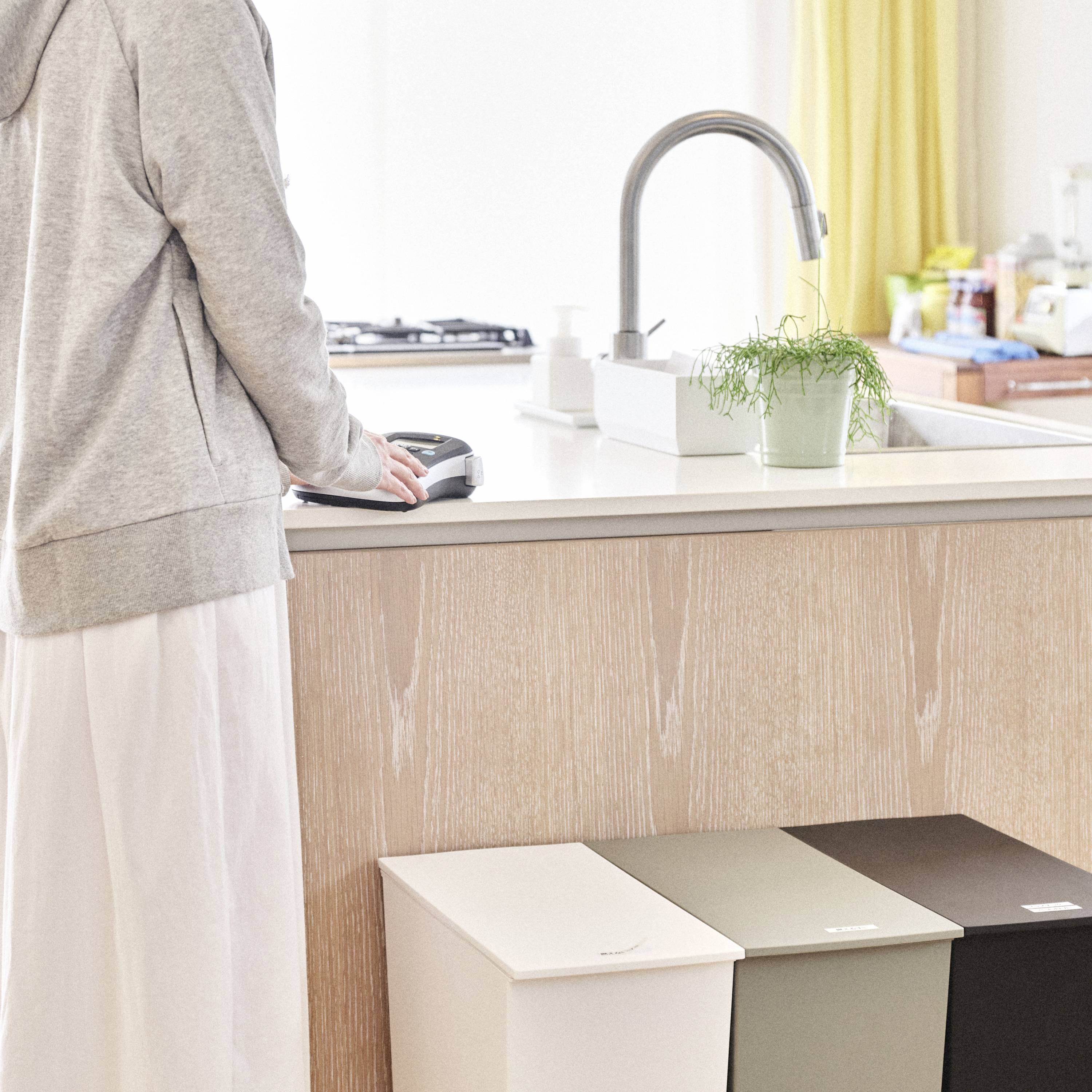 Eine Person steht an einer Küchentheke und spült Geschirr ab. Im Vordergrund sind drei Müllbehälter in verschiedenen Farben zu sehen.