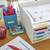 Ein Tisch mit Ordnern aufgestapelt, Etiketten: 'To Be Graded', 'Graded Papers', 'Exams', 'Quizzes', 'Homework'. Stifte und Büroklammern daneben.