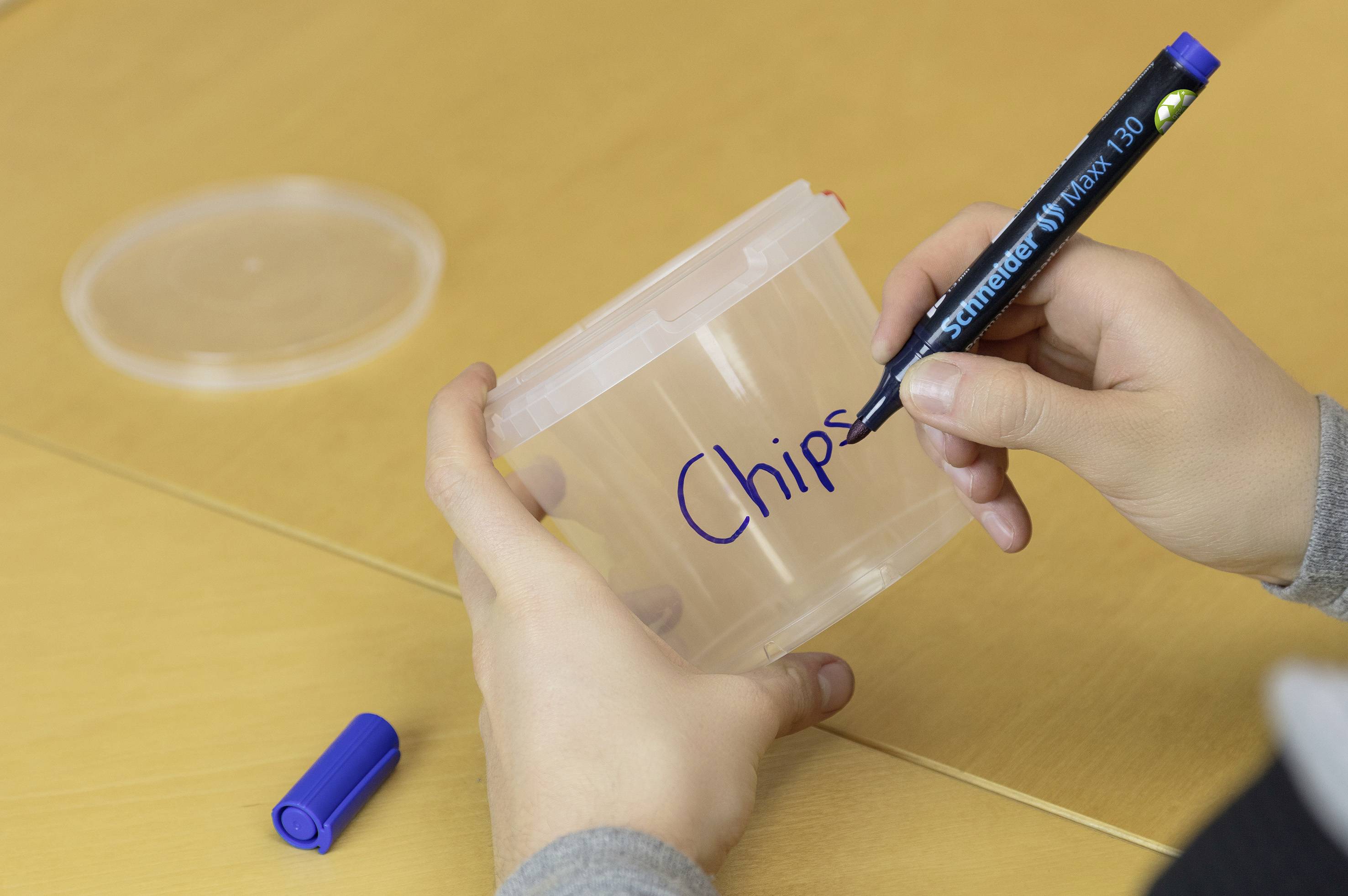 Eine Person beschriftet einen transparenten Plastikbehälter mit dem Wort 'Chips' mit einem blauen Permanentmarker. 
