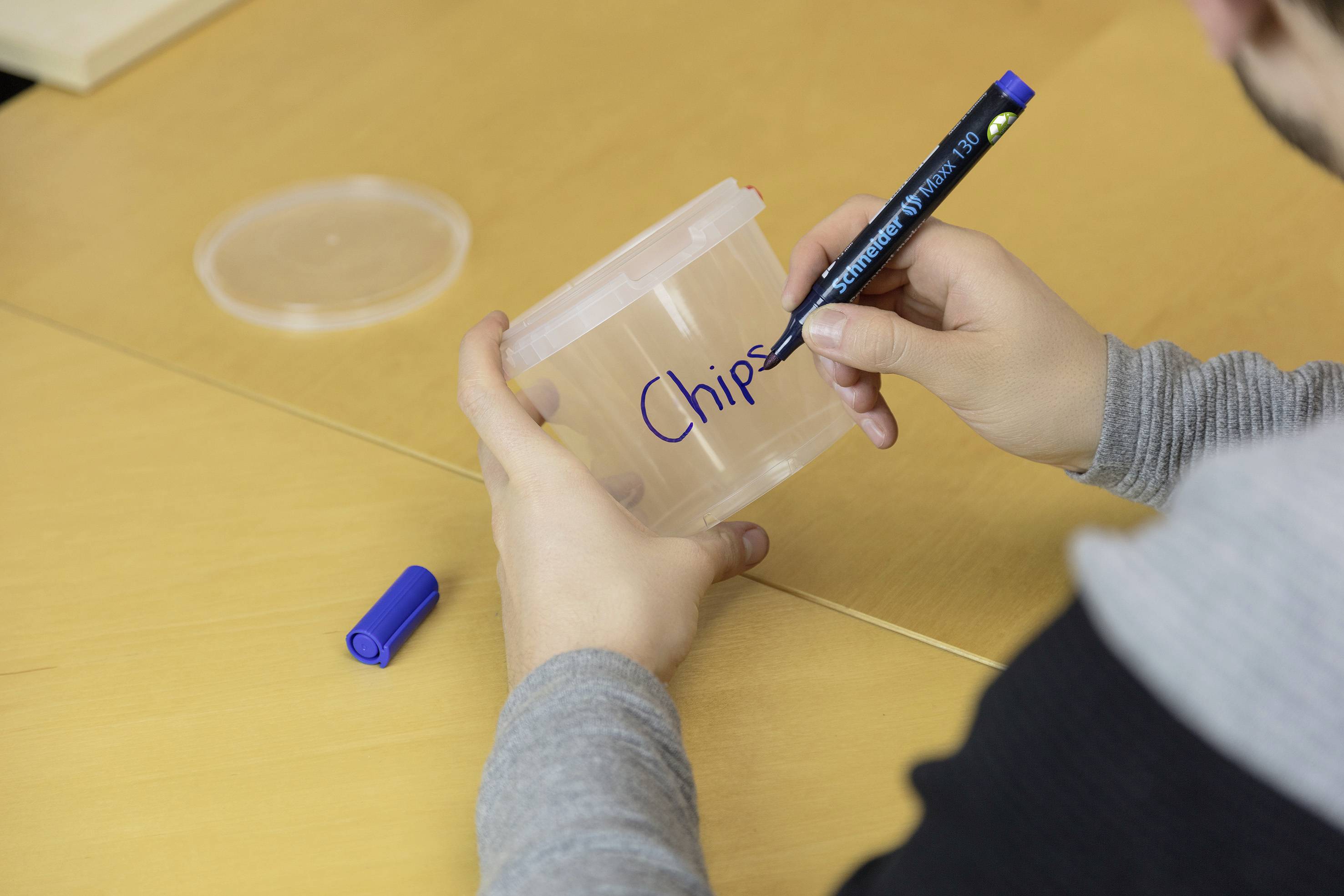 Eine Person beschriftet mit einem blauen Marker einen Plastikbehälter mit 'Chips'. Der Deckel liegt daneben auf einem Holztisch.