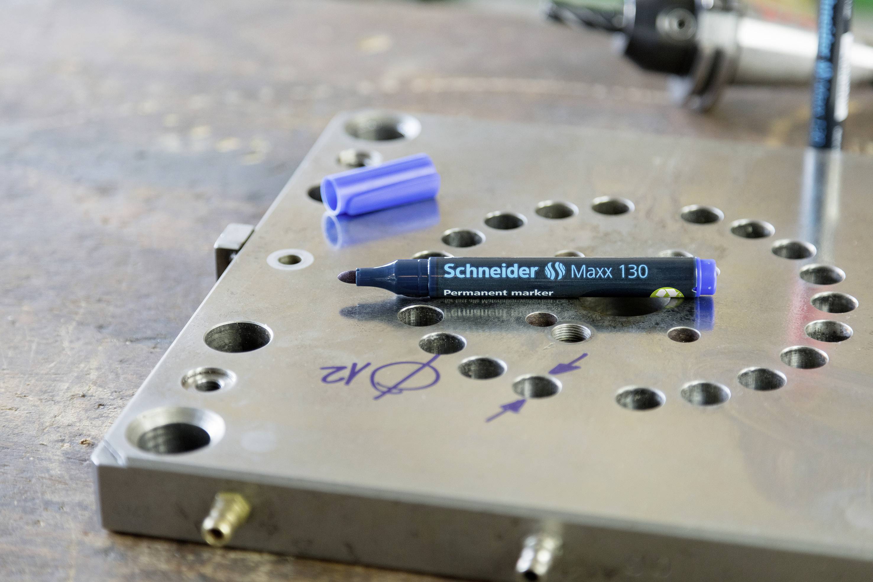 Metallplatte mit Löchern; ein blauer Permanentmarker liegt darauf. Im Hintergrund unscharfe Werkzeuge.