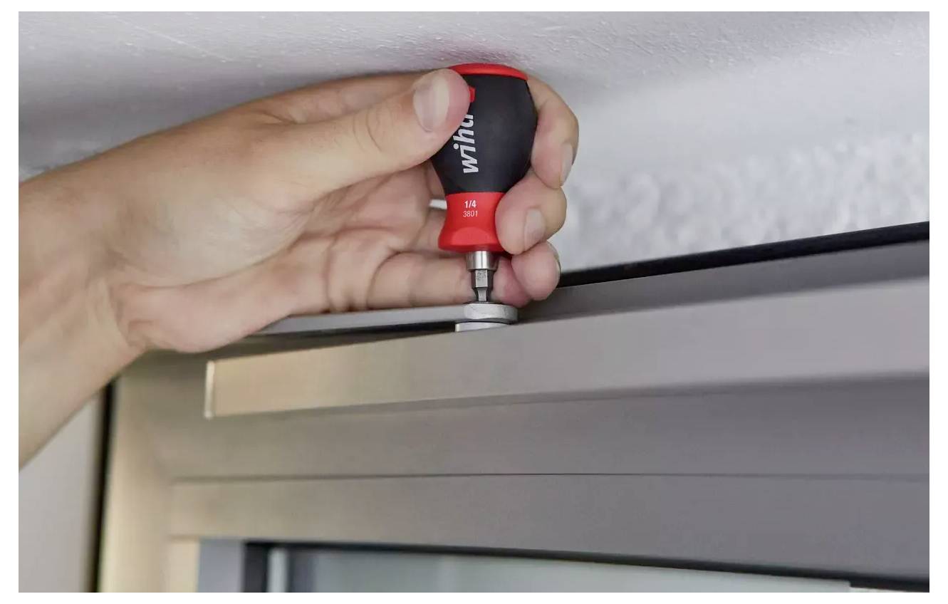 Eine Hand benutzt einen rot-schwarzen Schraubendreher, um ein Scharnier an einem Fensterrahmen einzustellen, was auf eine Heimwerker- oder Reparaturaufgabe hindeutet.