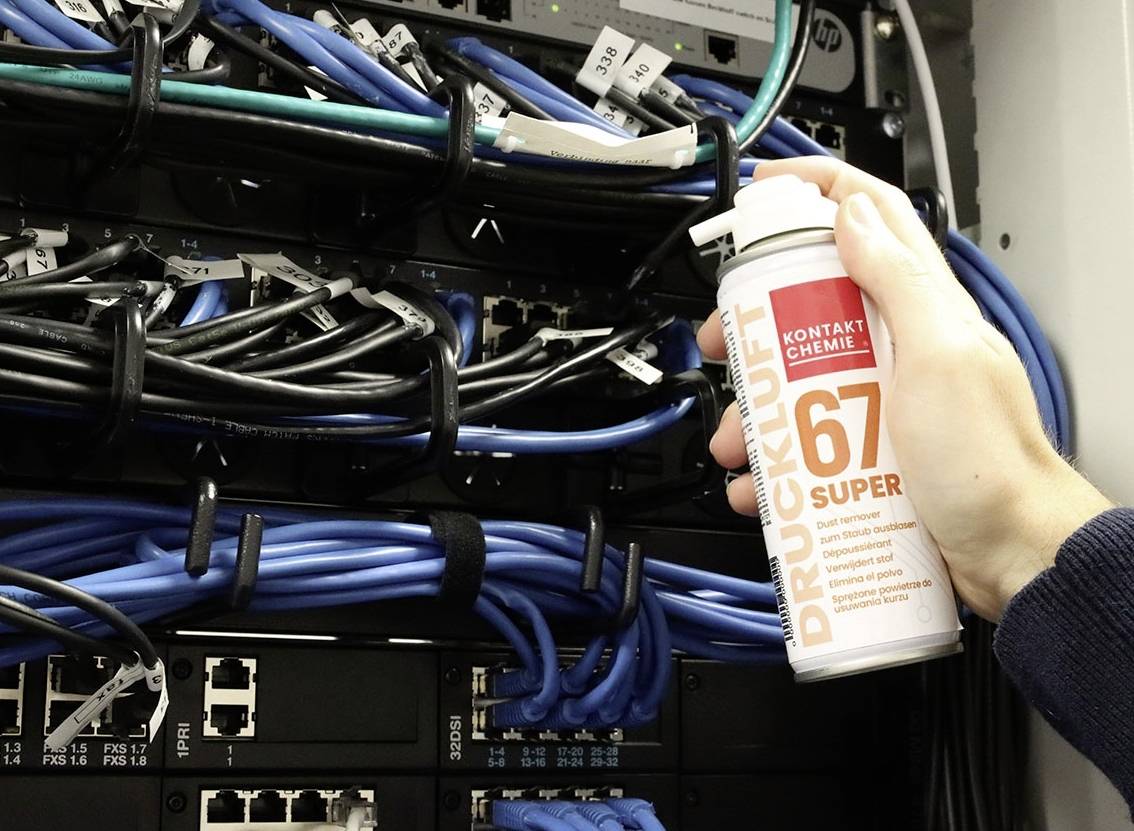 Eine Hand hält eine Dose Druckluftreiniger vor einem Netzwerk-Server. Kabel und Ports sind sichtbar.