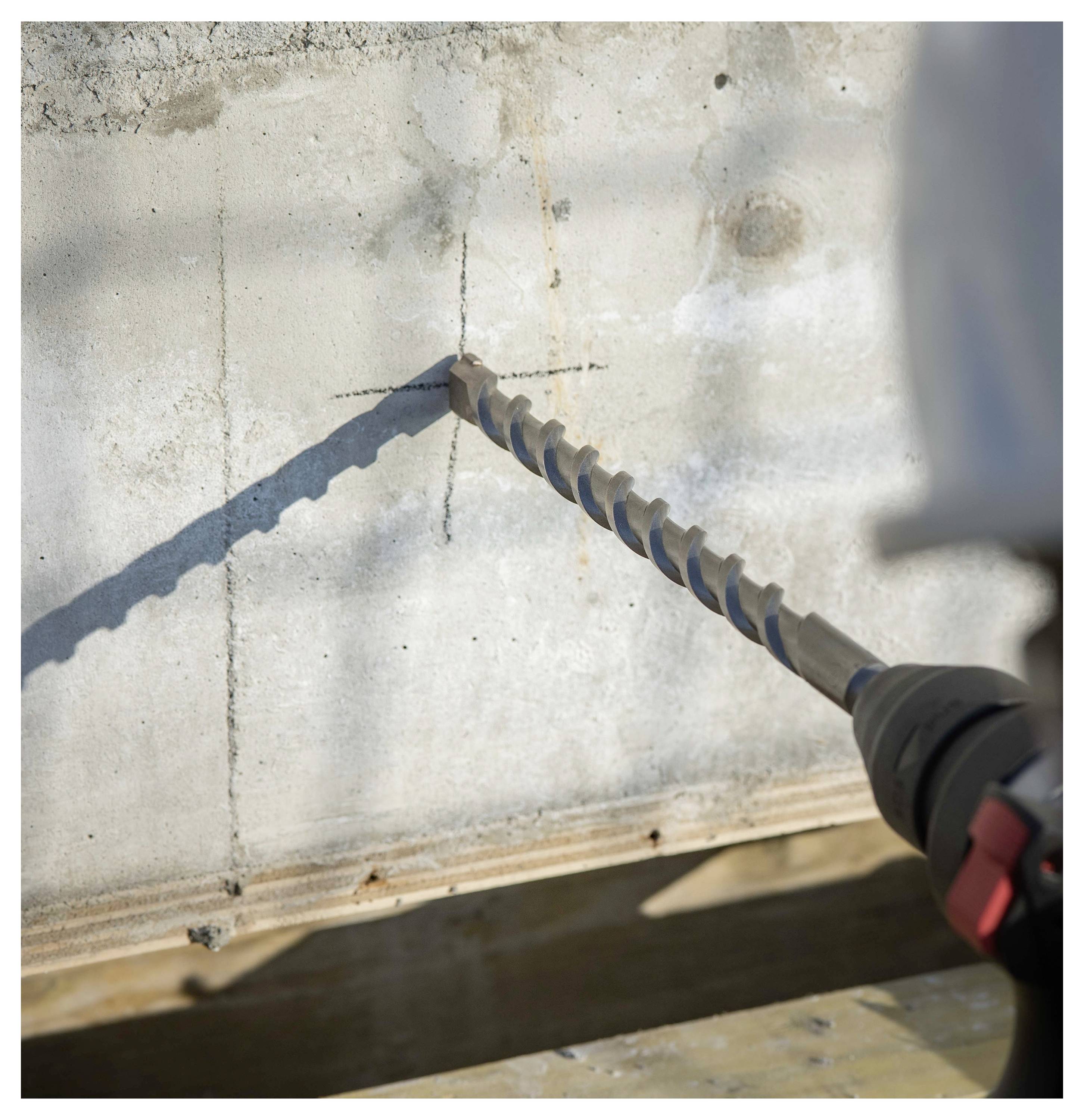 Ein Bohrer mit einem langen Steinbohrer bohrt in eine Betonwand, wobei ein markanter Bohrpunkt und das in einem Winkel gehaltene Werkzeug sichtbar sind.