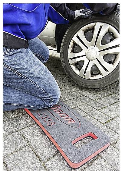 Eine Person kniet neben einem Autoreifen auf einer gepflasterten Oberfläche und verwendet eine Kniematte für Komfort, während sie den Reifen überprüft.