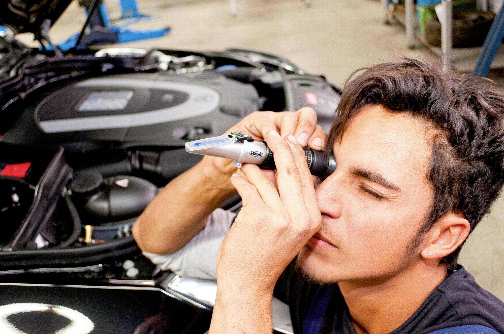 Ein Mechaniker benutzt ein Handgerät, um den Motor eines Autos zu inspizieren. Im Hintergrund sind Werkzeuge und eine Hebebühne sichtbar.