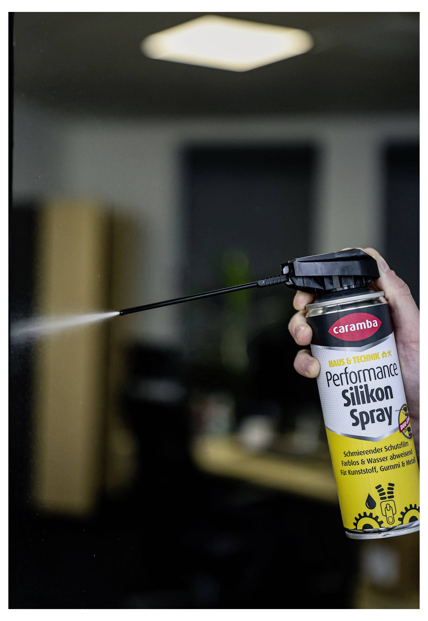 Manuelles Sprühen von Caramba Performance Silicon Spray aus einer Spraydose mit langer Düse in einer Werkstatt-Umgebung im Innenbereich.