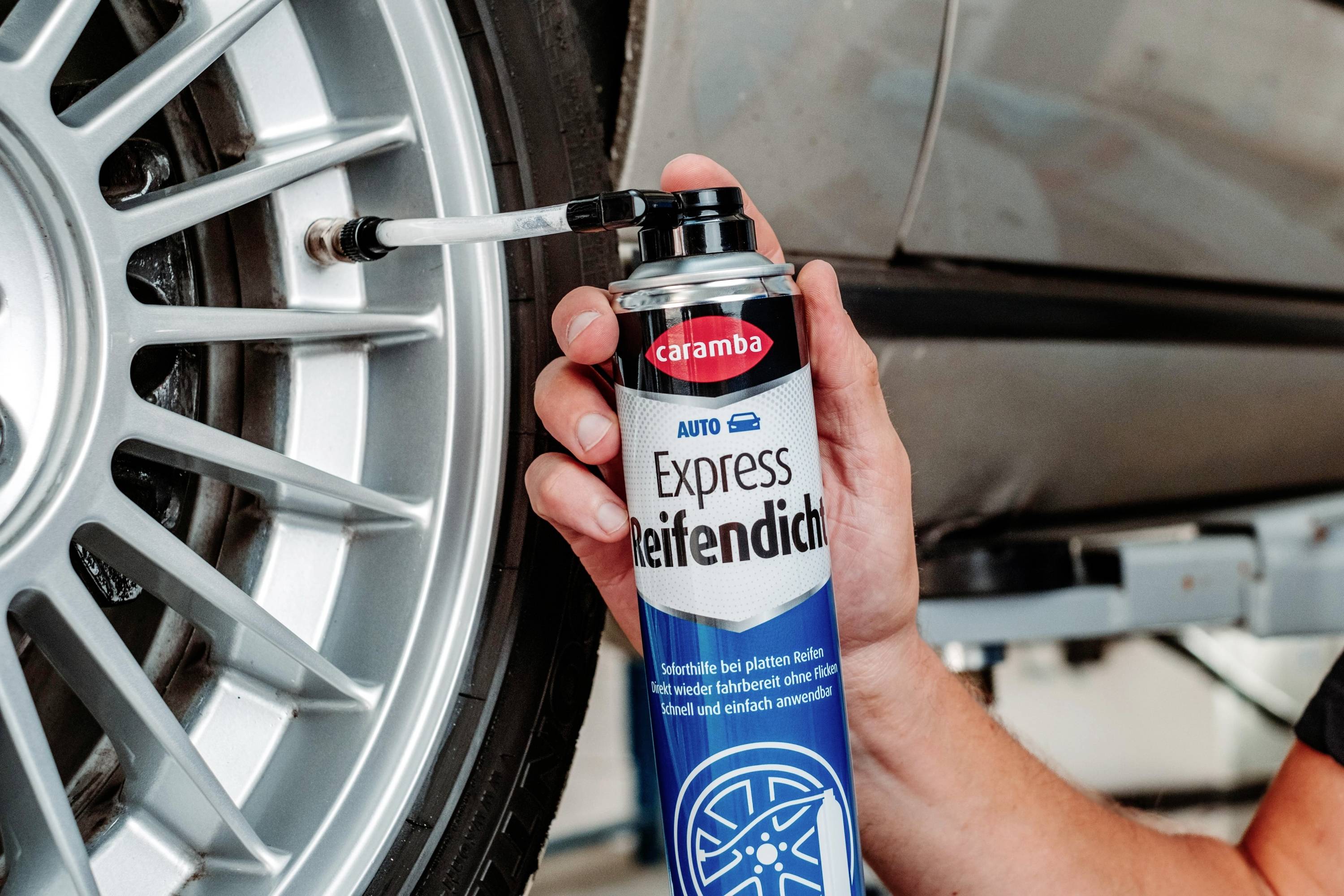 Person, die eine Dose 'Caramba Auto Express Reifendicht' Reifendichtmittel verwendet, um einen Reifen an einem Fahrzeug auf einer Hebebühne in einer Garagenumgebung zu reparieren.