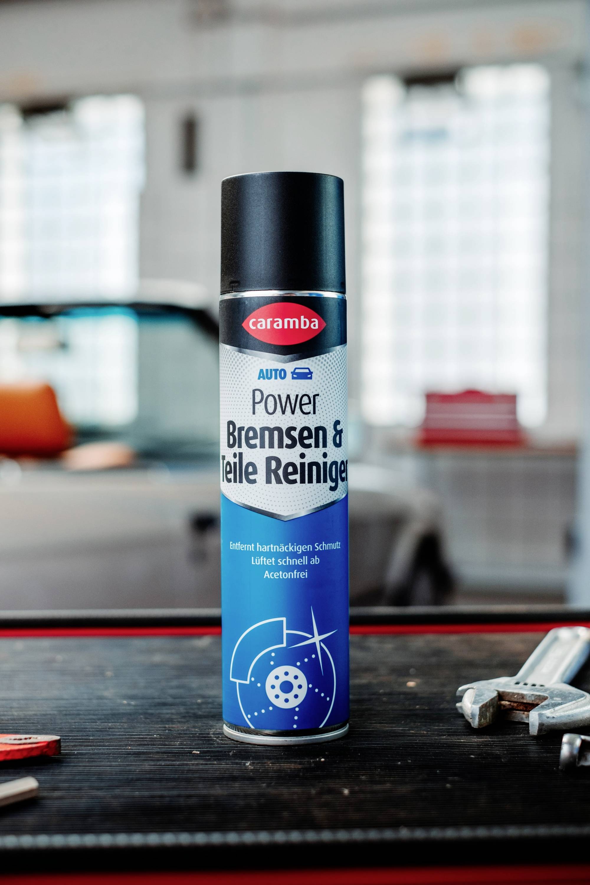 Eine Dose Caramba Power Bremsen & Teile Reiniger steht auf einer Werkbank, mit einem Auto im unscharfen Hintergrund.