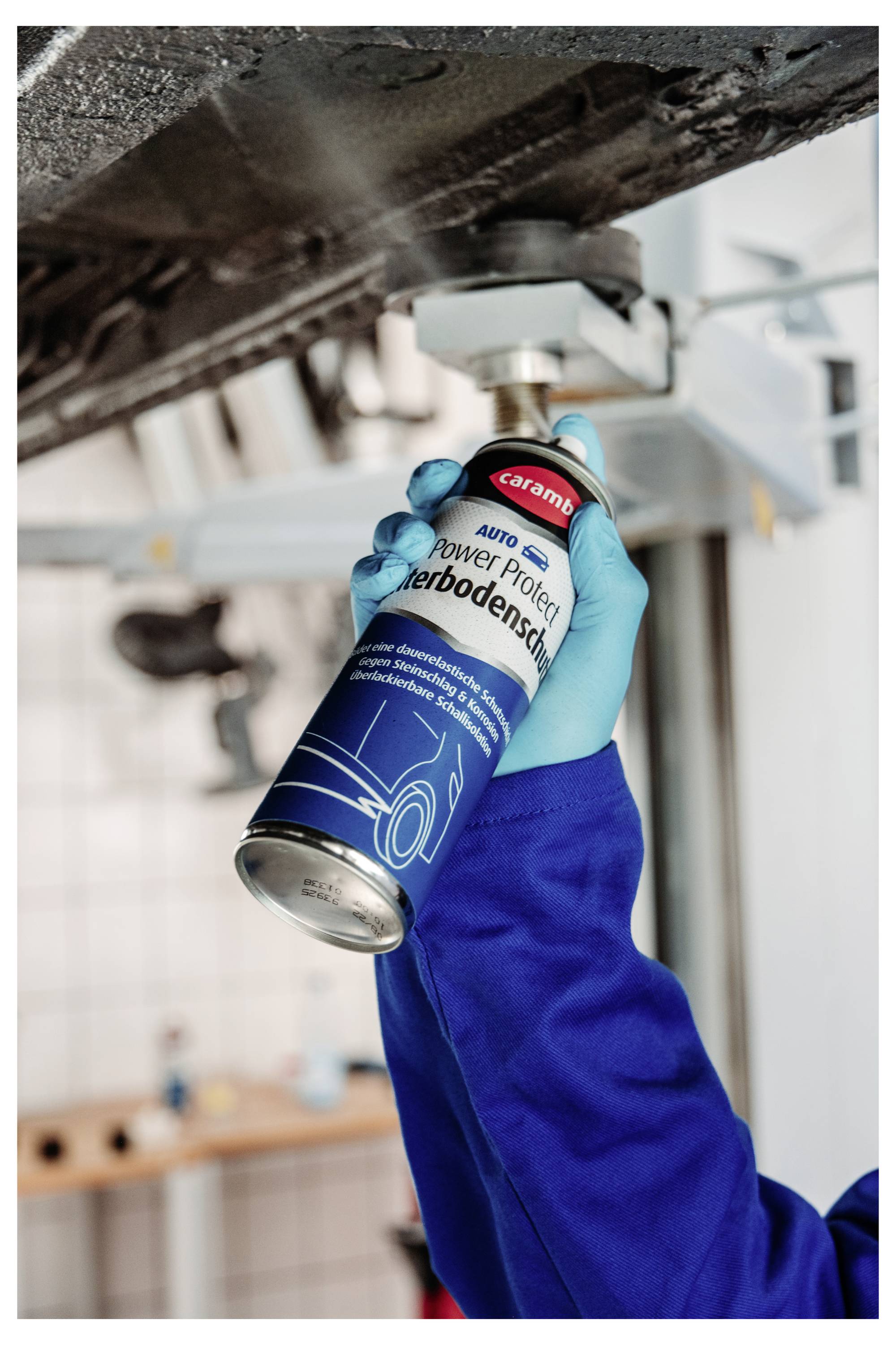 Arbeiter in blauen Handschuhen hält eine Sprühdose und trägt Unterbodenschutz oder Rostschutz auf die Unterseite eines Fahrzeugs auf einer Hebebühne auf.
