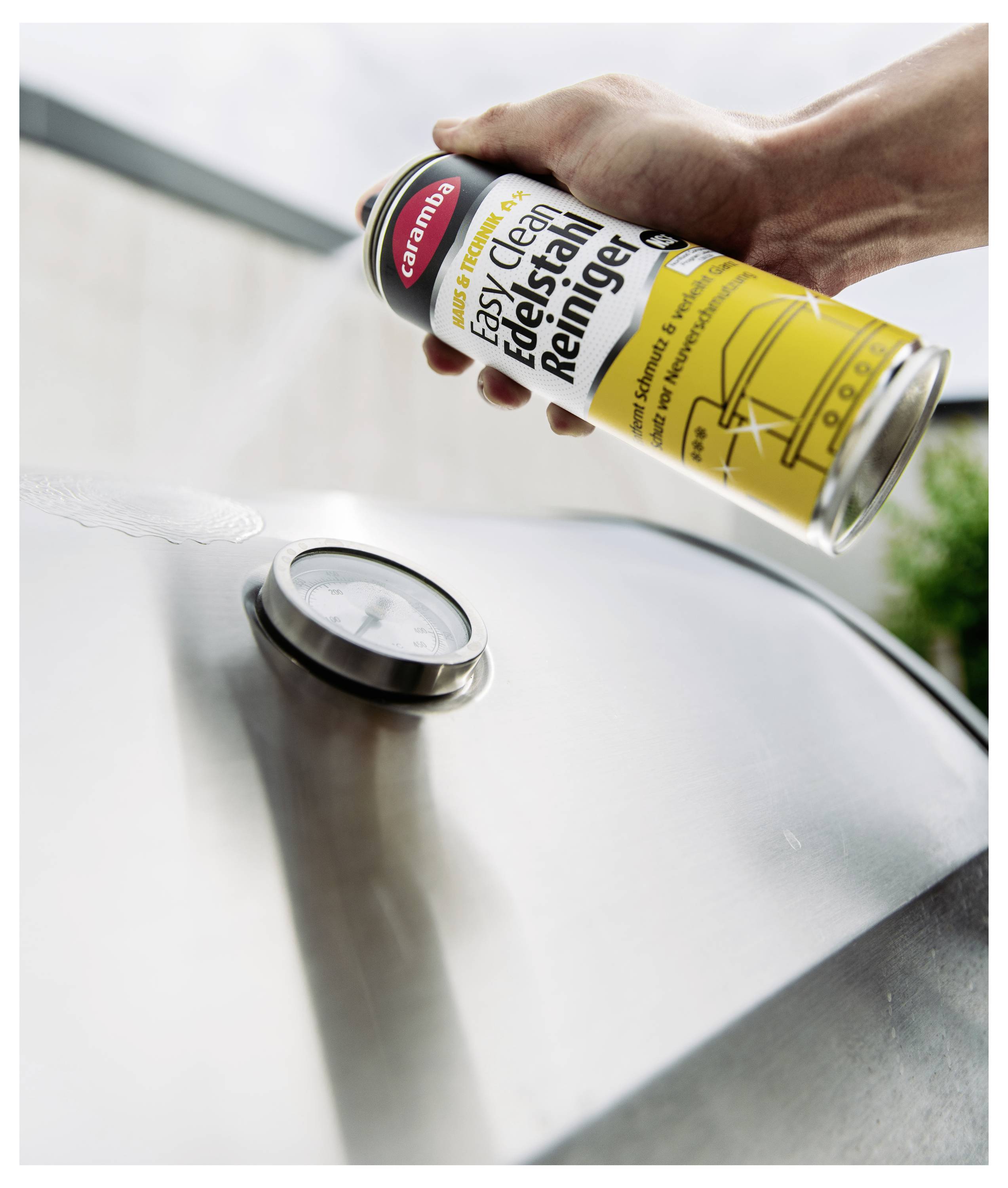 Hand hält eine Dose "easy clean Edelstahl Reiniger" über einen Edelstahl-Grilldeckel mit Thermometer, was auf die Reinigung von Geräten hindeutet.