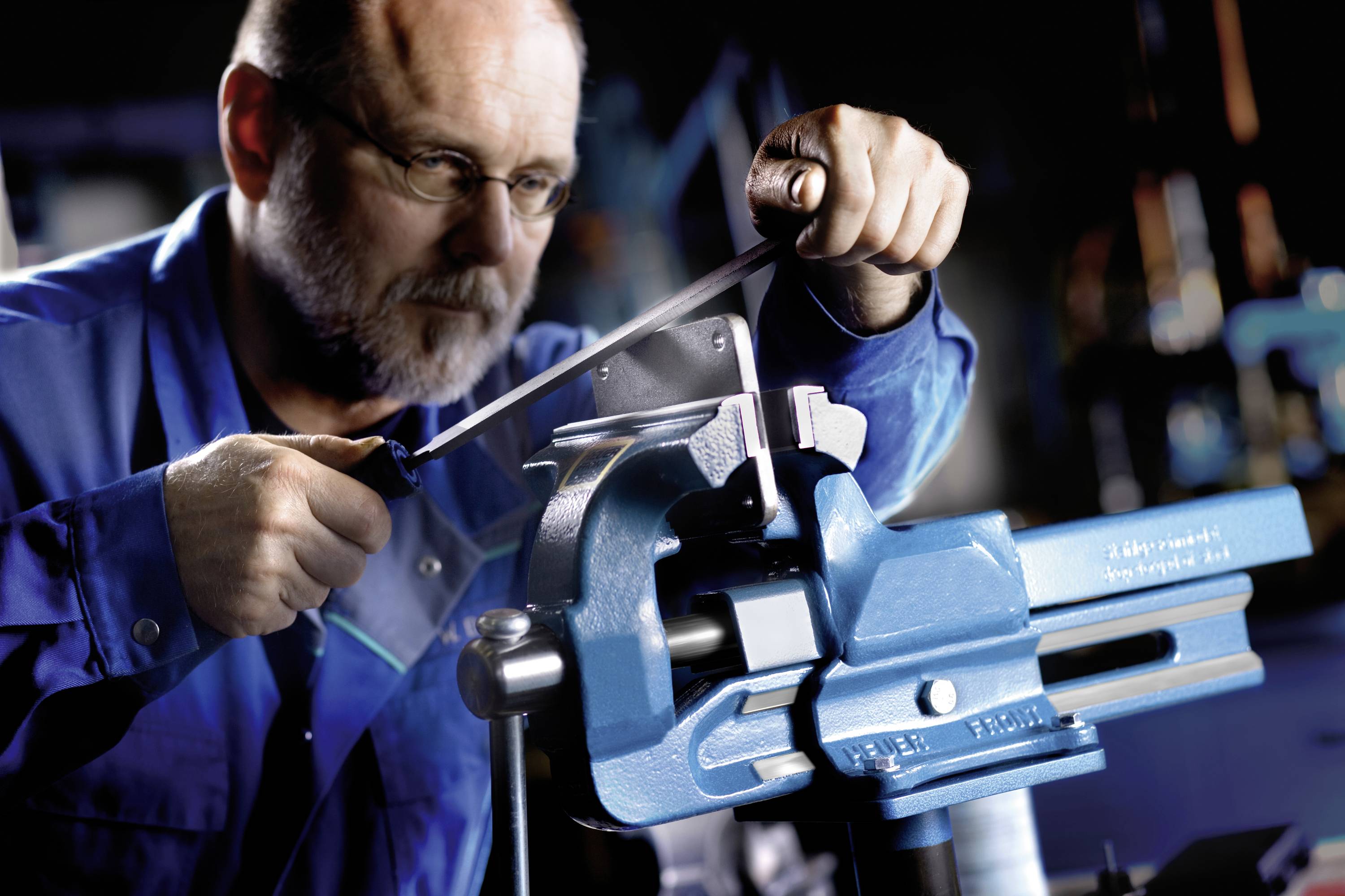 Ein Mann bearbeitet ein Metallstück mit einer Feile, das in einem blauen Schraubstock eingespannt ist.
