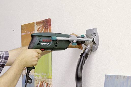 Person benutzt eine grüne Bohrmaschine an einer weißen Wand mit einem Absaugsystem, um sauberes Bohren zu ermöglichen.