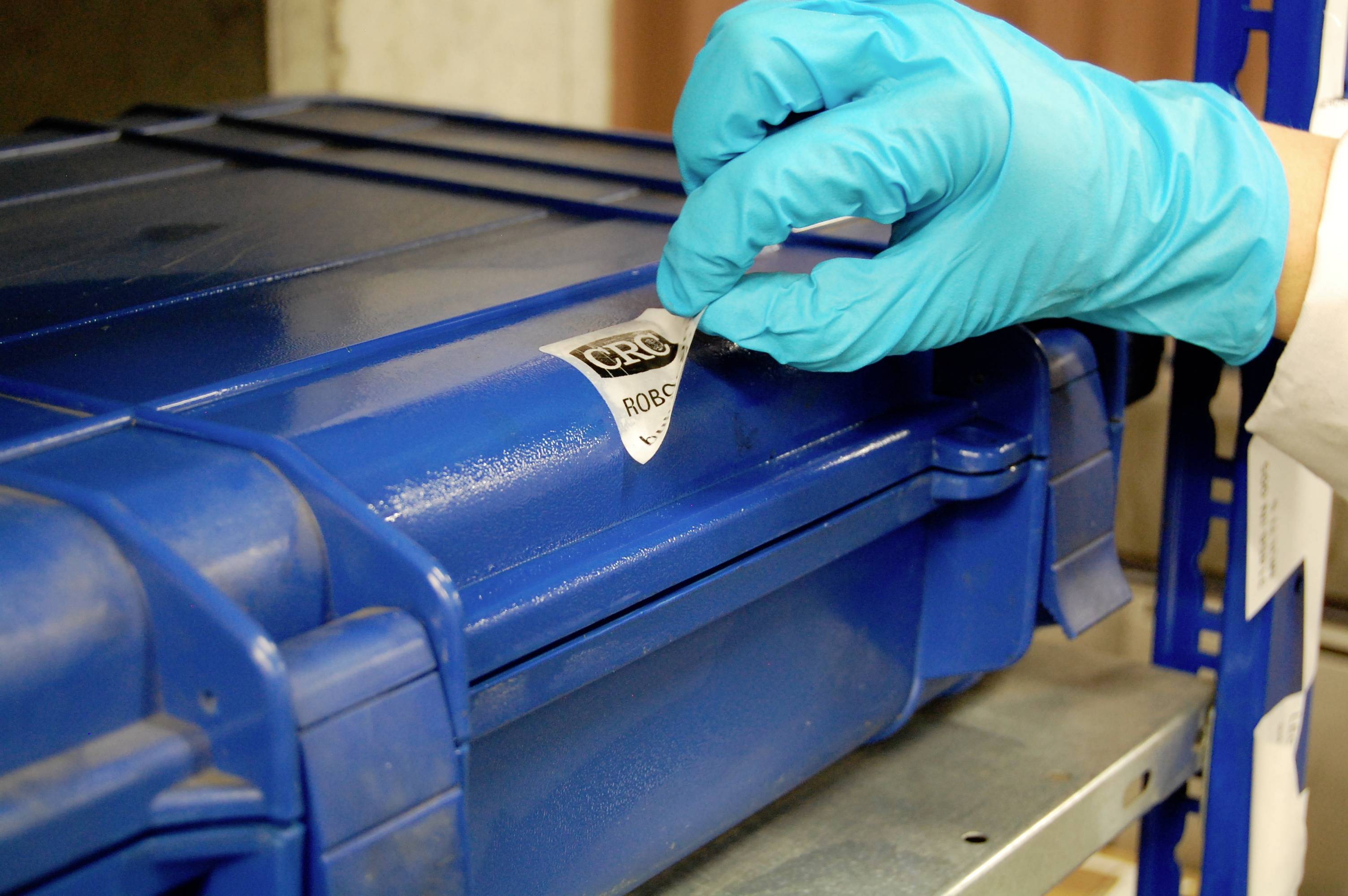 Eine behandschuhte Hand entfernt ein Sicherheitssiegel von einem blauen, verschlossenen Koffer. Der Koffer steht auf einem Regal.